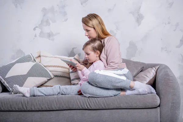 Beautiful young mother and her little daughter are watching movies together and playing on the tablet while sitting on the sofa. Mom hugs daughter. Maternal care and love. Horizontal photo