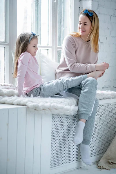 Hermosa madre joven y su hija pequeña están sentados junto a la ventana juntos y pintándose las uñas. Cuidado materno y amor. Foto vertical — Foto de Stock