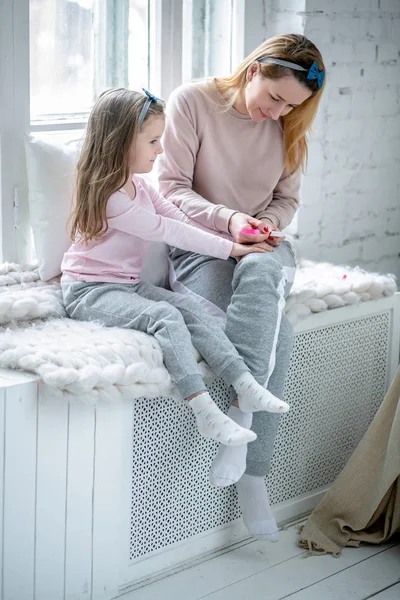 Hermosa madre joven y su hija pequeña están sentados junto a la ventana juntos y pintándose las uñas. Cuidado materno y amor. Foto vertical — Foto de Stock
