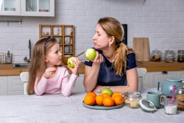 Mutlu küçük kız ve onun güzel genç annesi birlikte beyaz bir mutfakta kahvaltı var. Eğleniyorlar ve elma yiyorlar. Anne bakımı ve sevgi. Sağlıklı beslenme