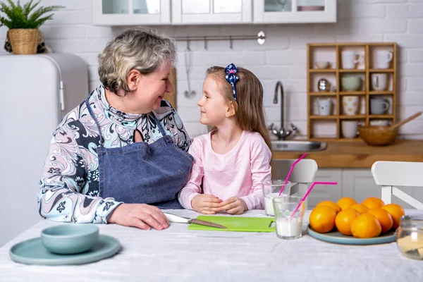 Happy little girl and her grandmother have breakfast together in a white kitchen. They are having fun and playing with fruits. Maternal care and love. Healthy eating