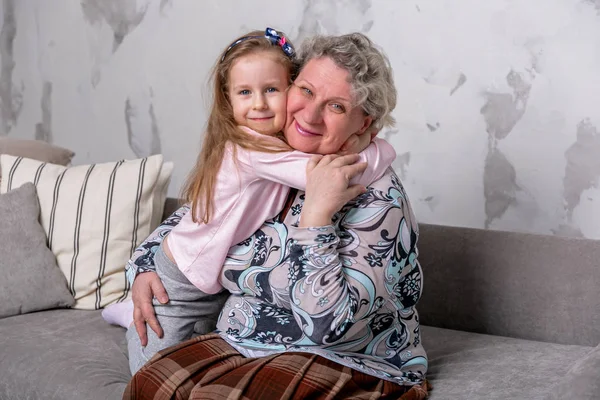 Grandmother and her little granddaughter are watching movies together and playing on the device while sitting on the sofa. Grandmom hugs granddaughter. Maternal care and love. Horizontal photo