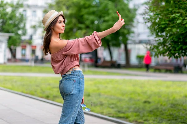 Fashionably dressed young woman taking selfie on the street on a sunny evening. Girl in jeans, blouse and small hat is looking at the display. Street-fashion concept. Late spring and early summer