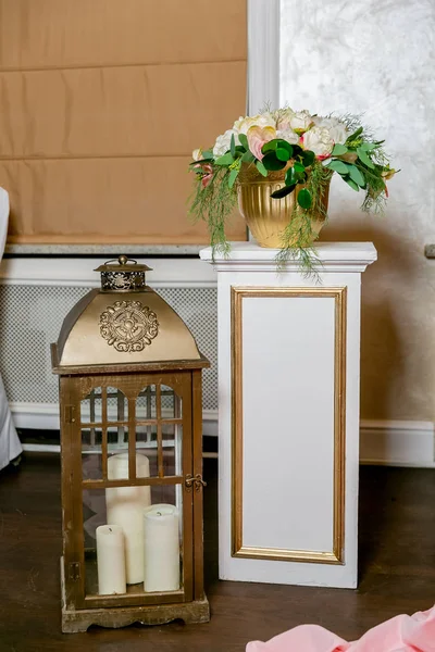 A white bouquet of peonies, hortensia and anthurium in a gold flowerpot on a white pillar in a classic style. Bronze lantern with candles on the floor