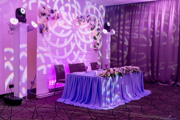 Wedding banquet in blue and white tones. Dinner table for newlyweds decorated with greenery and long cloth. A long flower arrangement on the table and a floral decor on a white wooden wall