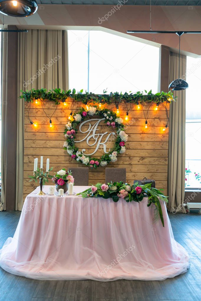 Beautiful white dinner table for newlyweds decorated with greenery and long cloth. A long flower arrangement on the table, a floral wreath with an emblem and a garland of light bulbs on a wooden wall