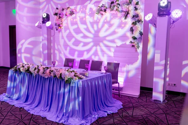 Wedding banquet in blue and white tones. Dinner table for newlyweds decorated with greenery and long cloth. A long flower arrangement on the table and a floral decor on a white wooden wall