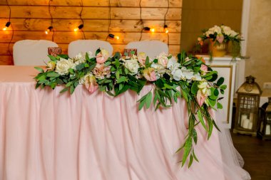 Yeşillik ve uzun bez ile dekore yeni evliler için güzel beyaz yemek masası. Masada uzun bir çiçek aranjmanı, çiçek kompozisyonu ve ahşap bir duvarda ampuller bir çelenk