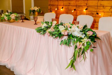 Yeşillik ve uzun bez ile dekore yeni evliler için güzel beyaz yemek masası. Masada uzun bir çiçek aranjmanı, çiçek kompozisyonu ve ahşap bir duvarda ampuller bir çelenk