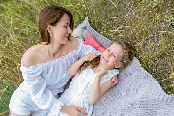 Krásná mladá matka a její dceruška v bílých šatech se baví na pikniku. Leží na plna trávy a smějí se. Mateřská péče a láska. Vodorovná fotografie — Stock fotografie