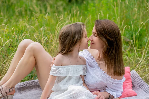 Beautiful young mother and her little daughter in white dress having fun in a picnic. They are sitting on a plaid on the grass and smiling. Maternal care and love. Horizontal photo — Stock Photo, Image