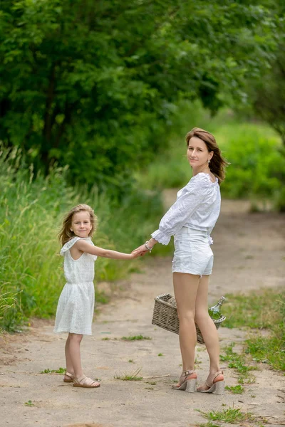 Bella giovane madre e la sua piccola figlia in abito bianco divertirsi in un pic-nic. Si fermano su una strada nel parco, si tengono per mano e guardano nella telecamera. Cura materna e amore. Foto verticale — Foto Stock