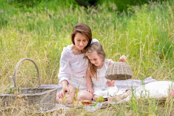 Тридцатилетняя красивая молодая мама и ее маленькая дочь в белом платье веселятся на пикнике. Они сидят на клетке на траве и достают сэндвичи. Материнская забота и любовь — стоковое фото