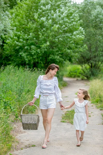 Красива молода мати і її маленька дочка в білій сукні розважаються на пікніку. Вони тримають руки і дивляться один на одного, ходять по дорозі в парку. Догляд за дітьми та кохання. Вертикальне фото — стокове фото