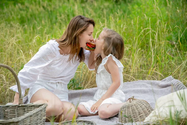 Krásná mladá matka a její dceruška v bílých šatech se baví na pikniku. Sedí na plání trávy a jedí sendvič společně. Mateřská péče a láska — Stock fotografie