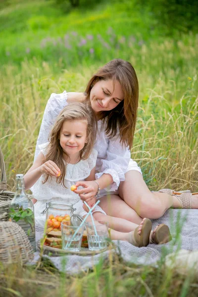 Красива молода мати і її маленька дочка в білій сукні розважаються на пікніку. Вони сидять на килимку і виймають ягоди з банки. Догляд за дітьми та кохання. Вертикальне фото — стокове фото