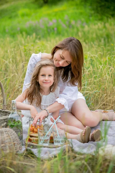 Beautiful young mother and her little daughter in white dress having fun in a picnic. They sit on the rug and take the berries out of the jar. Maternal care and love. Verticalal photo — Stock Photo, Image