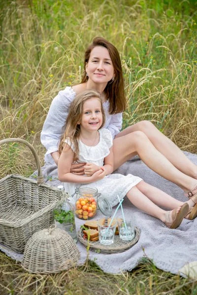 Красива молода мати і її маленька дочка в білій сукні розважаються на пікніку в літній день. Вони сидять на килимку і посміхаються. Догляд за дітьми та кохання. Вертикальне фото — стокове фото