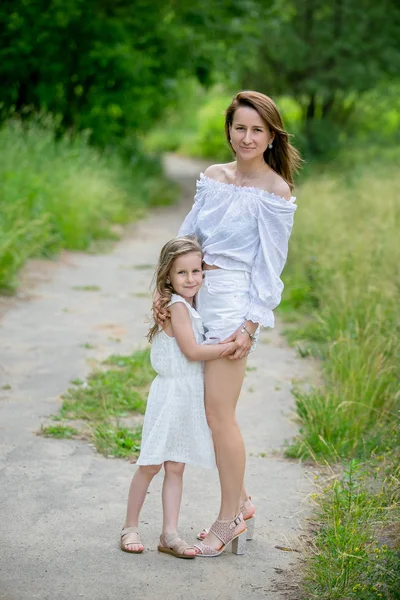 Krásná mladá matka a její dceruška v bílých šatech se baví na pikniku. Stojí na silnici v parku a objímají se. Mateřská péče a láska. Vertikální fotografie — Stock fotografie