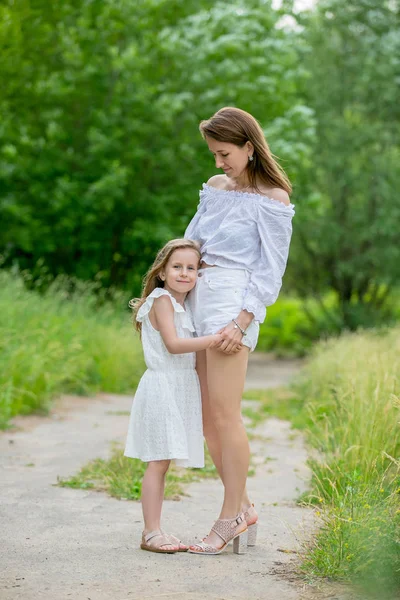 Schöne junge Mutter und ihre kleine Tochter im weißen Kleid haben Spaß bei einem Picknick. Sie stehen auf der Straße im Park und umarmen sich. Mütterfürsorge und Liebe. vertikales Foto — Stockfoto