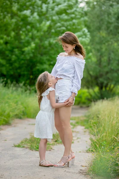 Beautiful young mother and her little daughter in white dress having fun in a picnic. They stand on the road in the park, hug and look at each other. Maternal care and love. Vertical photo — Stock Photo, Image