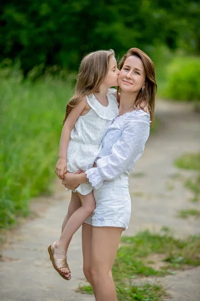 Krásná mladá matka a její dceruška v bílých šatech se baví na pikniku. Stojí na silnici v parku, Máma drží dceru v náručí, dcera líbá mámu. Mateřská péče a láska — Stock fotografie