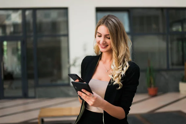Beautiful blonde business woman looking at tablet and phone, standing inside a company where she is working.
