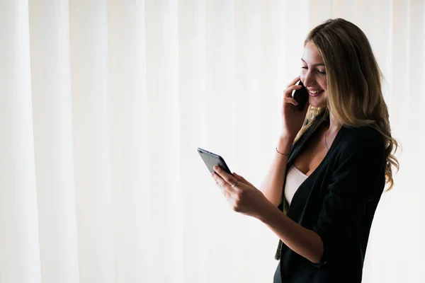 Young successful blonde business woman talking on phone in the office and smiling, looking at a tablet.