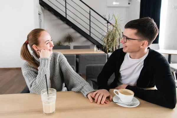 Foto av lyckliga unga paret hand i hand, tittar på varandra och ler medan du sitter på café — Stockfoto