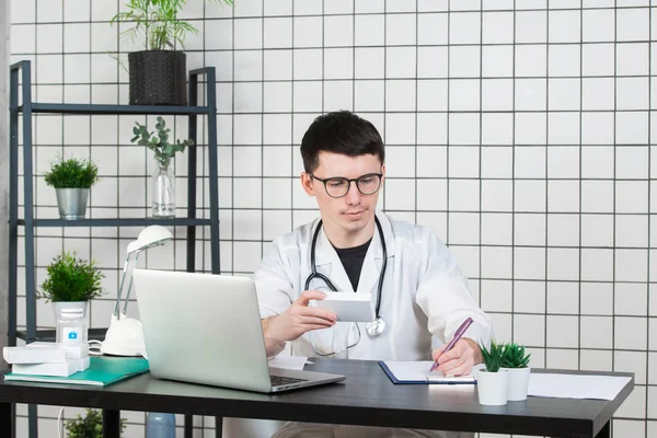 남자 의사 처방전에 생각 하는 테이블에 앉아 그의 목에 청진 기를 가진 백색 외 투에 적어 뭔가, 그의 손에서 의학의 상자 — 스톡 사진