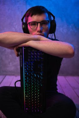 Portrait of the young handsome pro gamer sitting on the floor with keyboard in neon colored room clipart