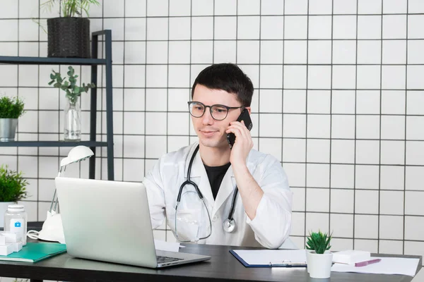 Male doctor using telephone while working on computer at table in clinic