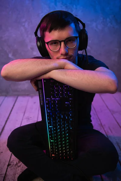 Porträt des jungen, gut aussehenden Profispielers, der mit Tastatur im neonfarbenen Raum auf dem Boden sitzt — Stockfoto