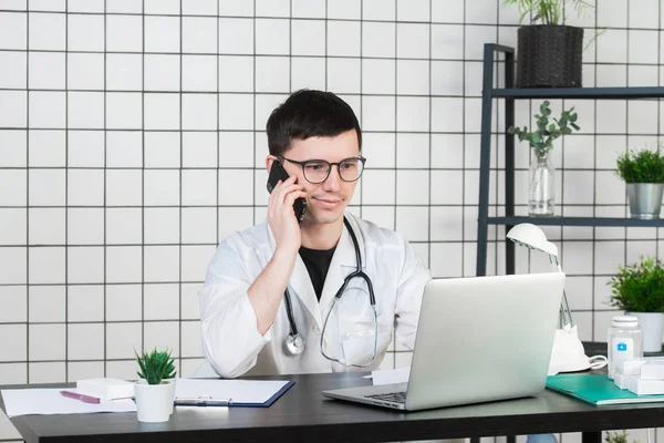 남자 의사 클리닉에 테이블에 컴퓨터에서 작업 하는 동안 전화를 사용 하 여 — 스톡 사진