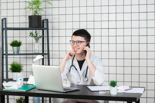 남자 의사 클리닉에 테이블에 컴퓨터에서 작업 하는 동안 전화를 사용 하 여 — 스톡 사진