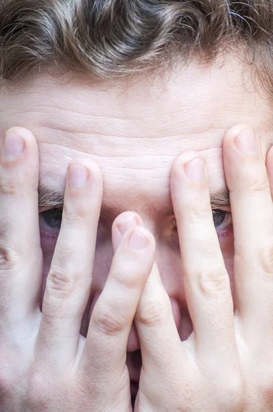 Portrait of young scared man hiding his eyes by covering his face with palms on white background.