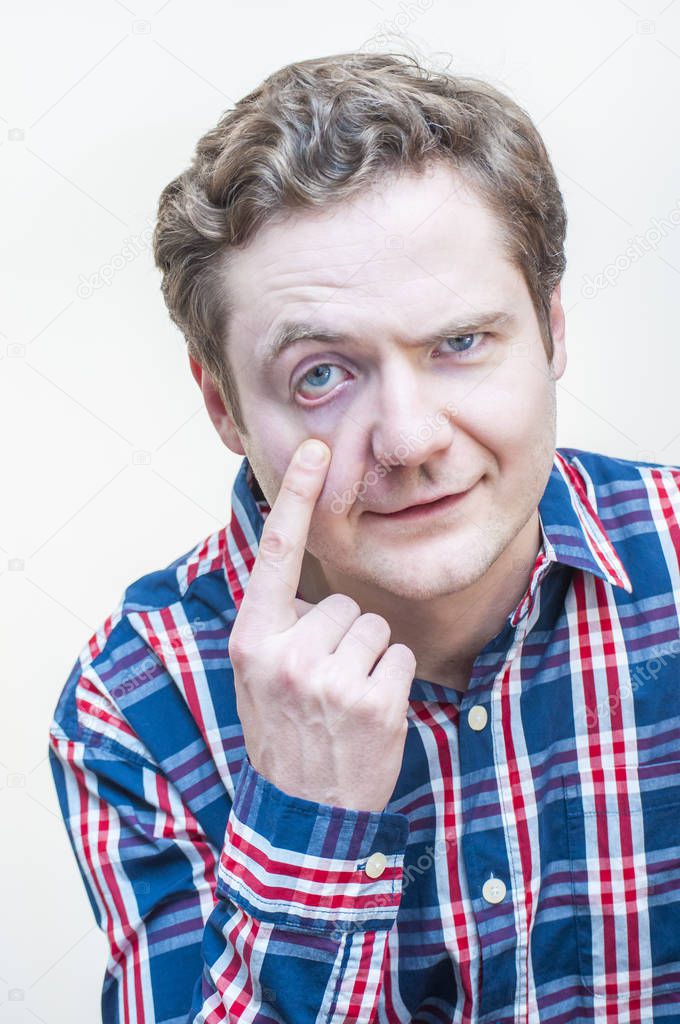 Portrait of young man questionably mistrustfully looking with raised eyebrow and opening down the same eye with his finger on white background.