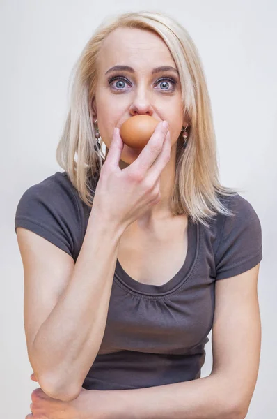 Big eyes young blonde woman holding chicken egg near her mouth on white background