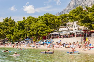 Makarska, Hırvatistan - 21 Haziran 2014: Yaz Makarska Plajı turistlerle dolu portresi. Ön plan, kıyı şeridi insanlar, çam ağaçları, restoranlar ve barlarla dolu buzlu deniz.