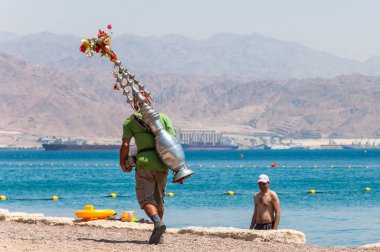 Eilat, İsrail - 15 Mayıs 2012: turist plajda Eilat, İsrail için sıcak bir içecek sunan adamla kırmızı şapka bakım otantik çay gemi konteyner sırtında,