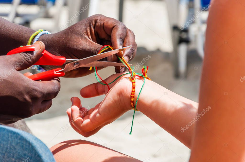 African ethnicity man cutting an extra threads of just braided bracelet from colored threads with scissors on caucasian ethnicity mans wrist on the beach. Seasonal beach resorts businesses