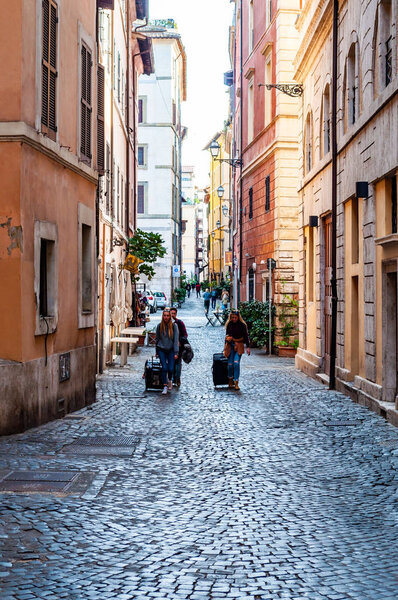 Rome, Italy - November 18, 2018: People walking with their luggage by the ancient medieval Old Town paving stone street in Rome, Italy