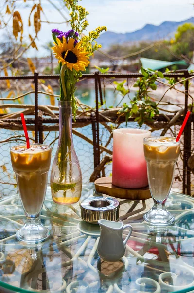 Two Ice coffee glasses with red drinking straw, vase with flowers, ashtray and red candle on the metal glass table surrounded by exotic plants. Romantic place.