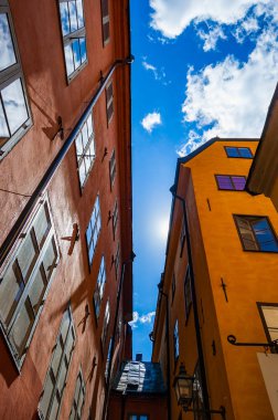 Aşağıdan rahat dar Ortaçağ sokak ile bağlı sarı turuncu kırmızı binalar cepheler Gamla stan, Stockholm eski kasaba parlak mavi gökyüzü ve güneş ışınları arka planda görüntüleyin