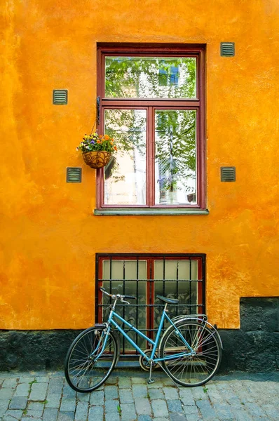 Azure vintage bicycle standing in front of vibrant orange building facade wall with big window with red window frame surrounded by several ventilation grills and hanging pot with blooming flowers