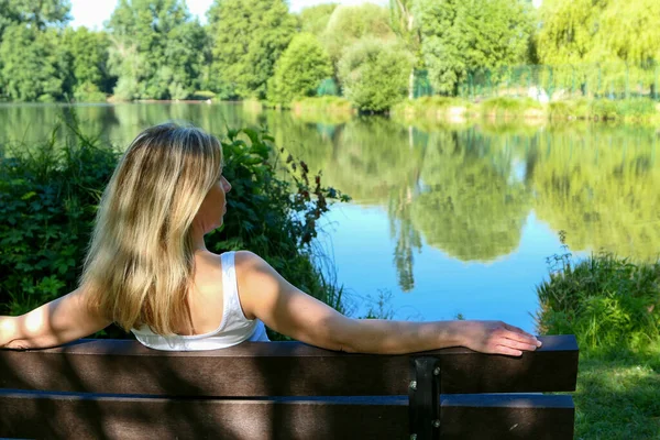 Blonde woman sitting on a bench in rural scene facing a lake in summer. Lonely female person relaxing in peace in nature. Deliberately blurred background.