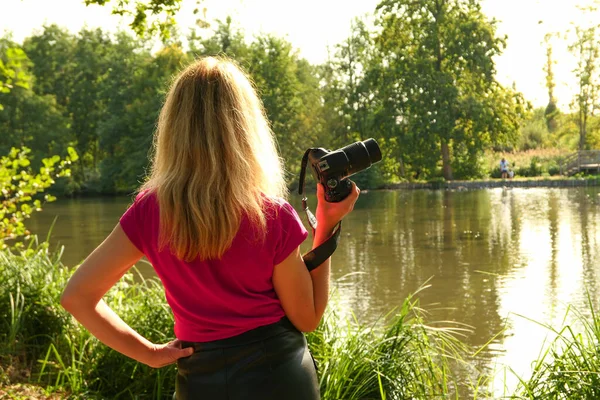 Blonde woman by a lake with a camera. Photographer in landscape with vegetation at sunrise.