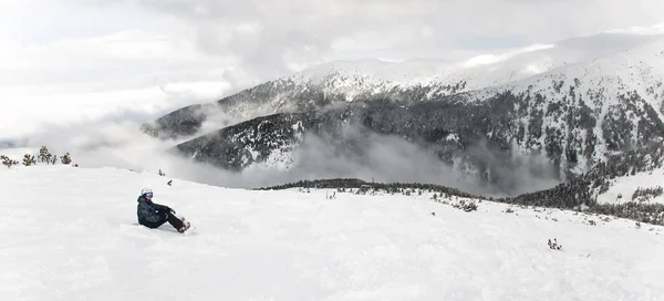 Snowboarder assis avec snowboard dans les mains est assis sur un gros rocher sur fond de montagnes. Bansko, Bulgarie — Photo