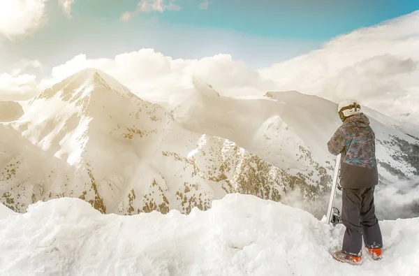 Snowboarder rester avec snowboard dans les mains est assis sur un gros rocher sur fond de montagnes. Bansko, Bulgarie — Photo