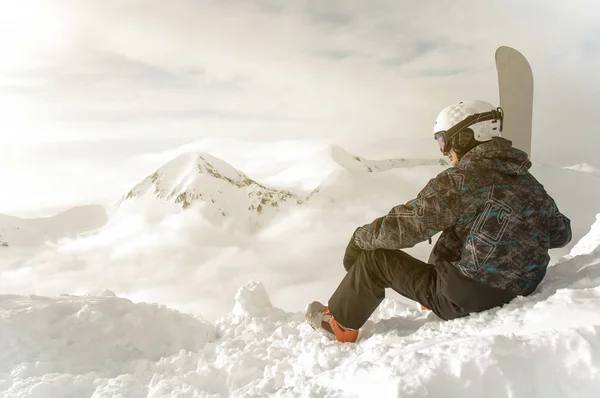 Snowboarder assis avec snowboard dans les mains est assis sur un gros rocher sur fond de montagnes. Bansko, Bulgarie — Photo